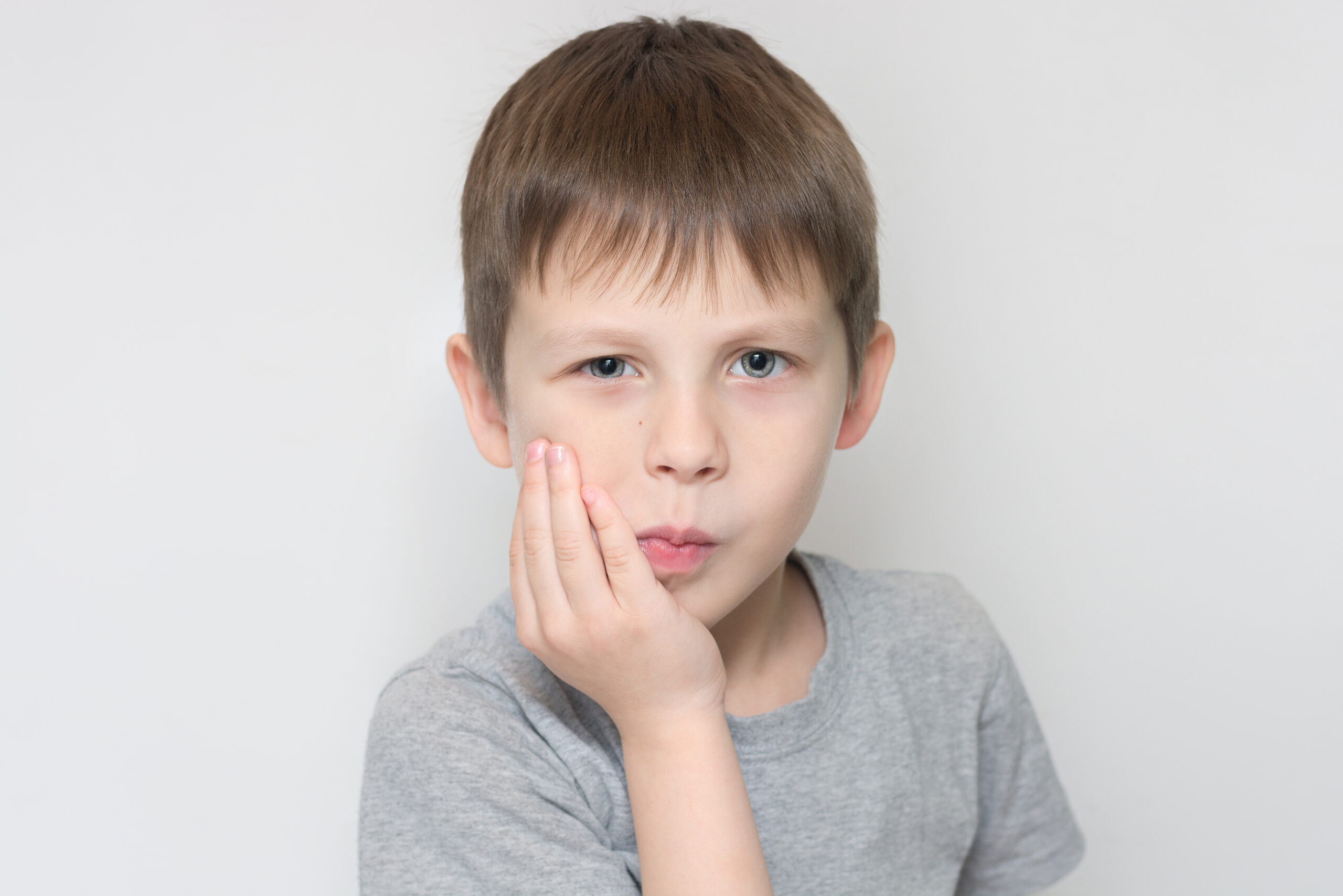 the child holds his cheek with his hand, indicating that a tooth hurts. The boy looks at the camera, horizontal photo, portrait. Unhappy preschooler with toothache